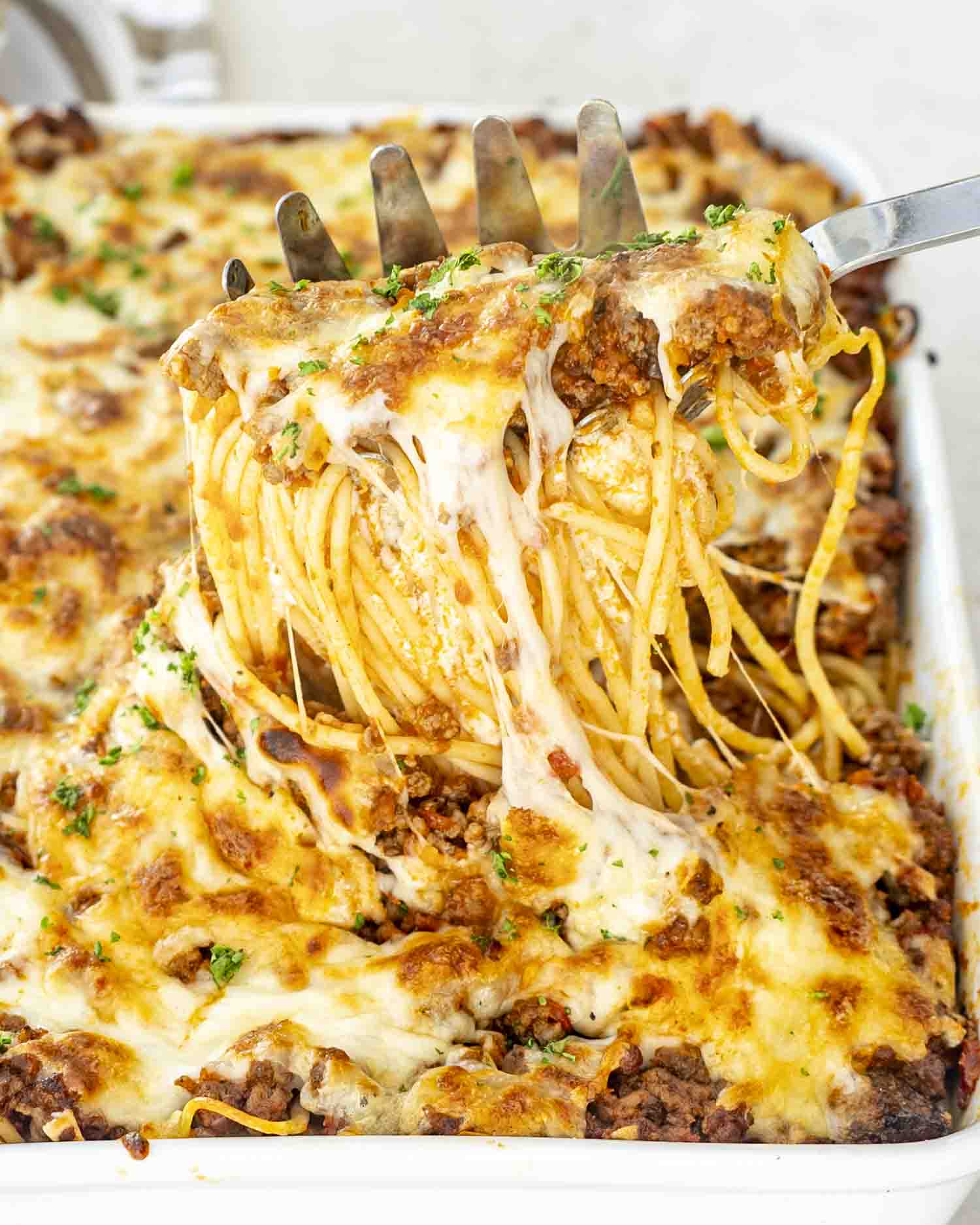 Golden, bubbly spaghetti slice being lifted from a cheesy baked casserole dish with stretchy mozzarella, rich meat sauce, and herby garnish.
