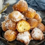 Close-up of golden ricotta zeppole stacked high on a plate, dusted with powdered sugar, with one cut open revealing the airy center.