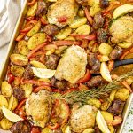 Sheet pan loaded with baked chicken, sausage, potatoes, peppers, lemon wedges, and fresh rosemary sprig.