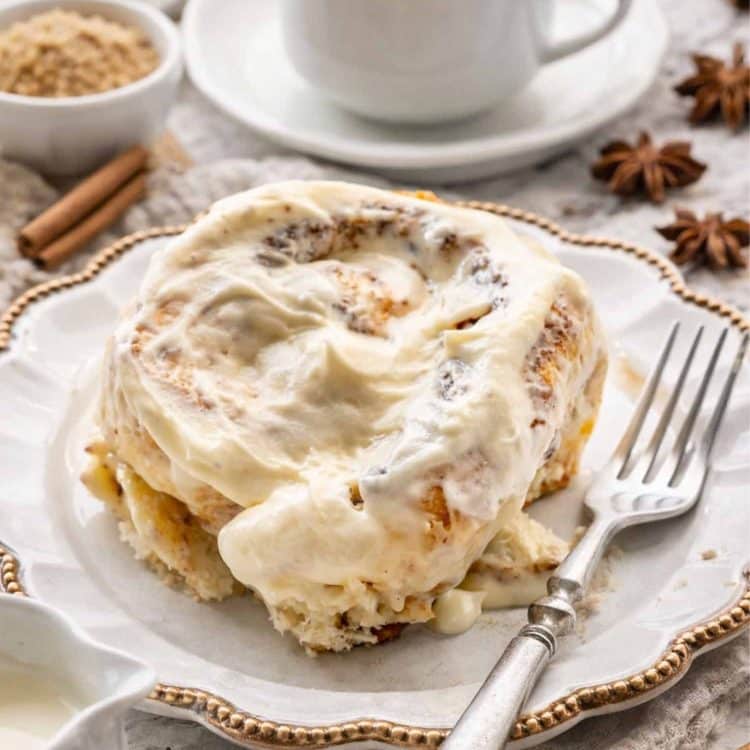 a cinnabon cinnamon roll on a white dessert plate with a cup of coffee in the background.
