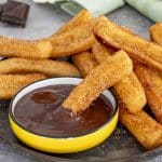 Cinnamon sugar churro dipped halfway into a bowl of rich melted chocolate, with more churros stacked beside it.