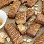 Rows of homemade Snickers Bites on parchment, coated in chocolate and caramel drizzle, with peanuts scattered around for texture.