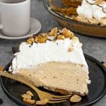 Creamy peanut butter pie slice on black plate with golden fork, garnished with peanuts and chocolate drizzle.