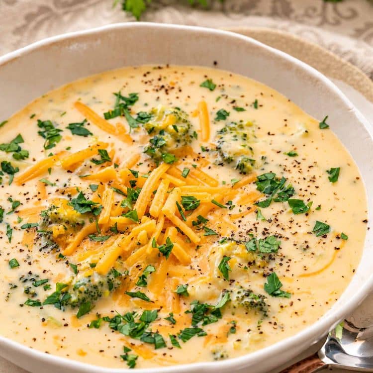 Close-up of creamy broccoli cheddar soup in a ceramic bowl, topped with shredded cheddar, chopped parsley, and black pepper.