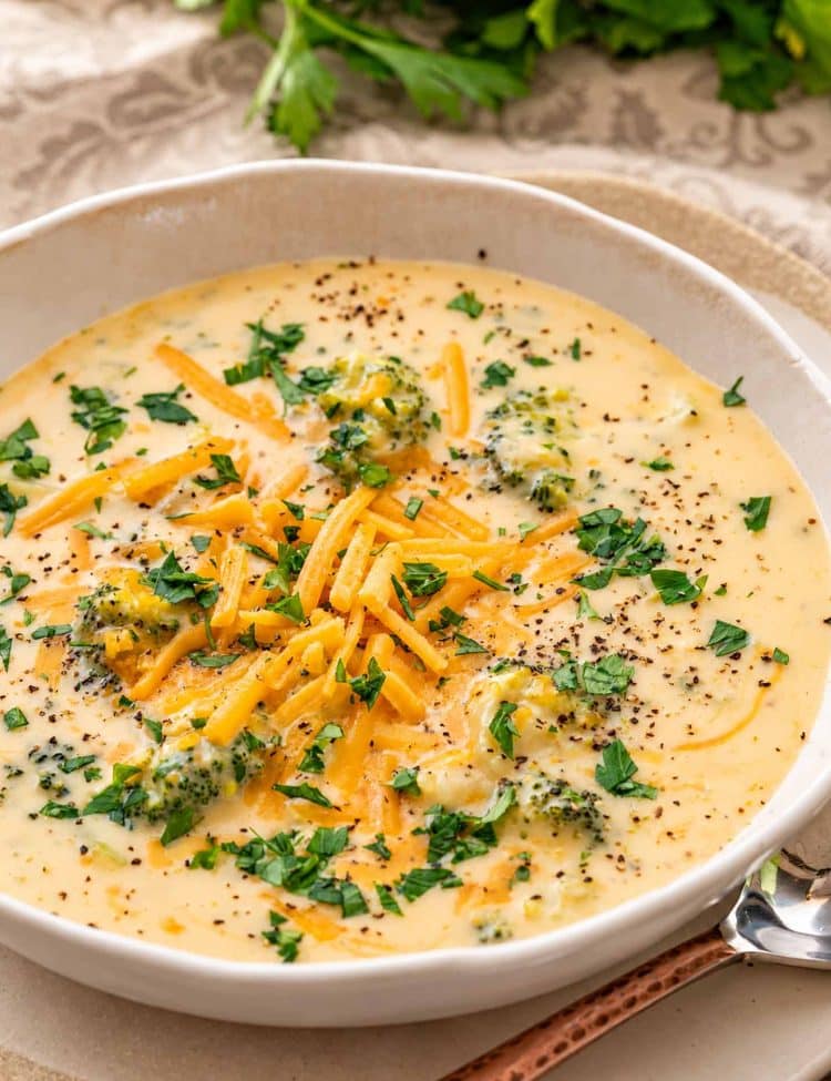 Close-up of creamy broccoli cheddar soup in a ceramic bowl, topped with shredded cheddar, chopped parsley, and black pepper.