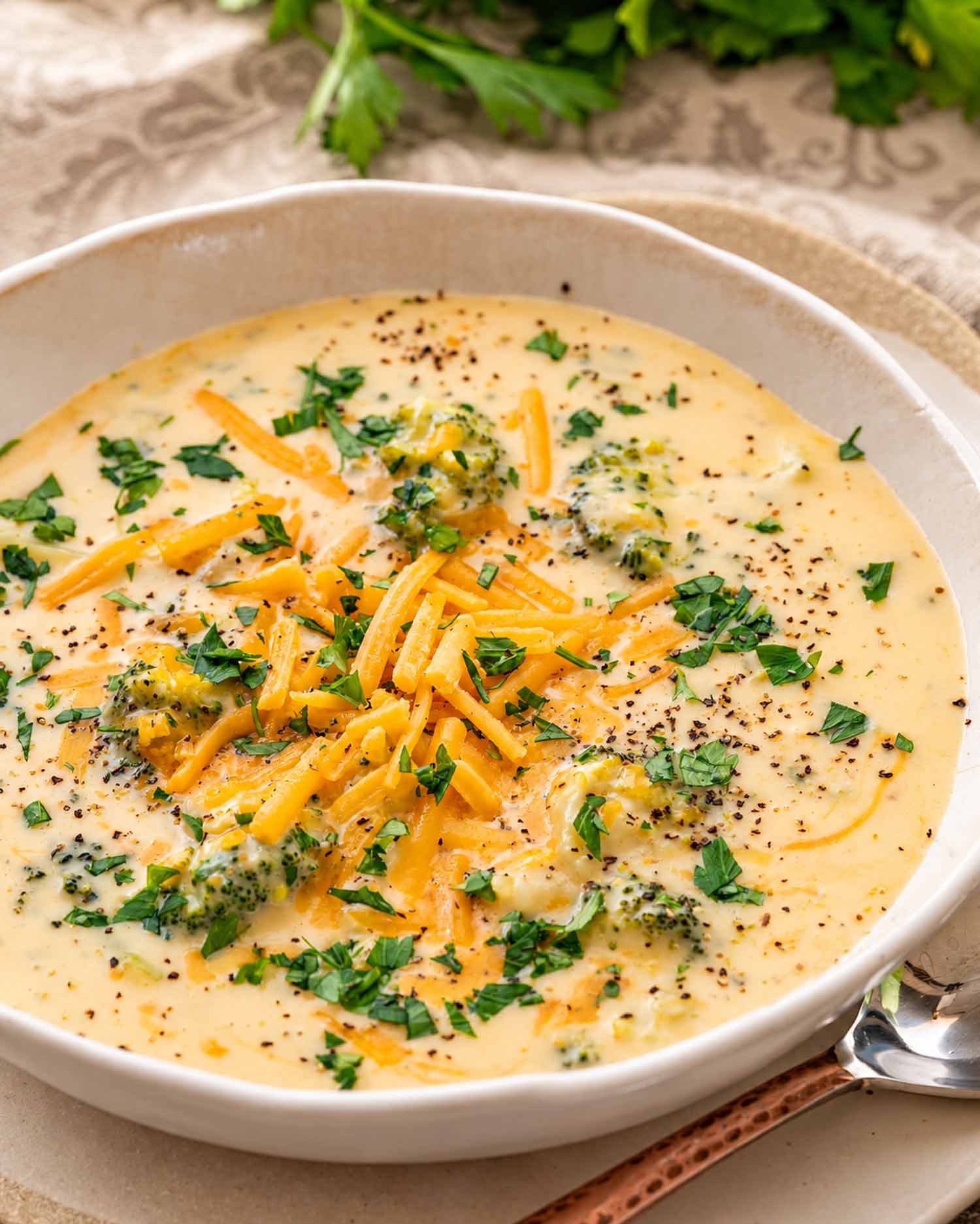Close-up of creamy broccoli cheddar soup in a ceramic bowl, topped with shredded cheddar, chopped parsley, and black pepper.