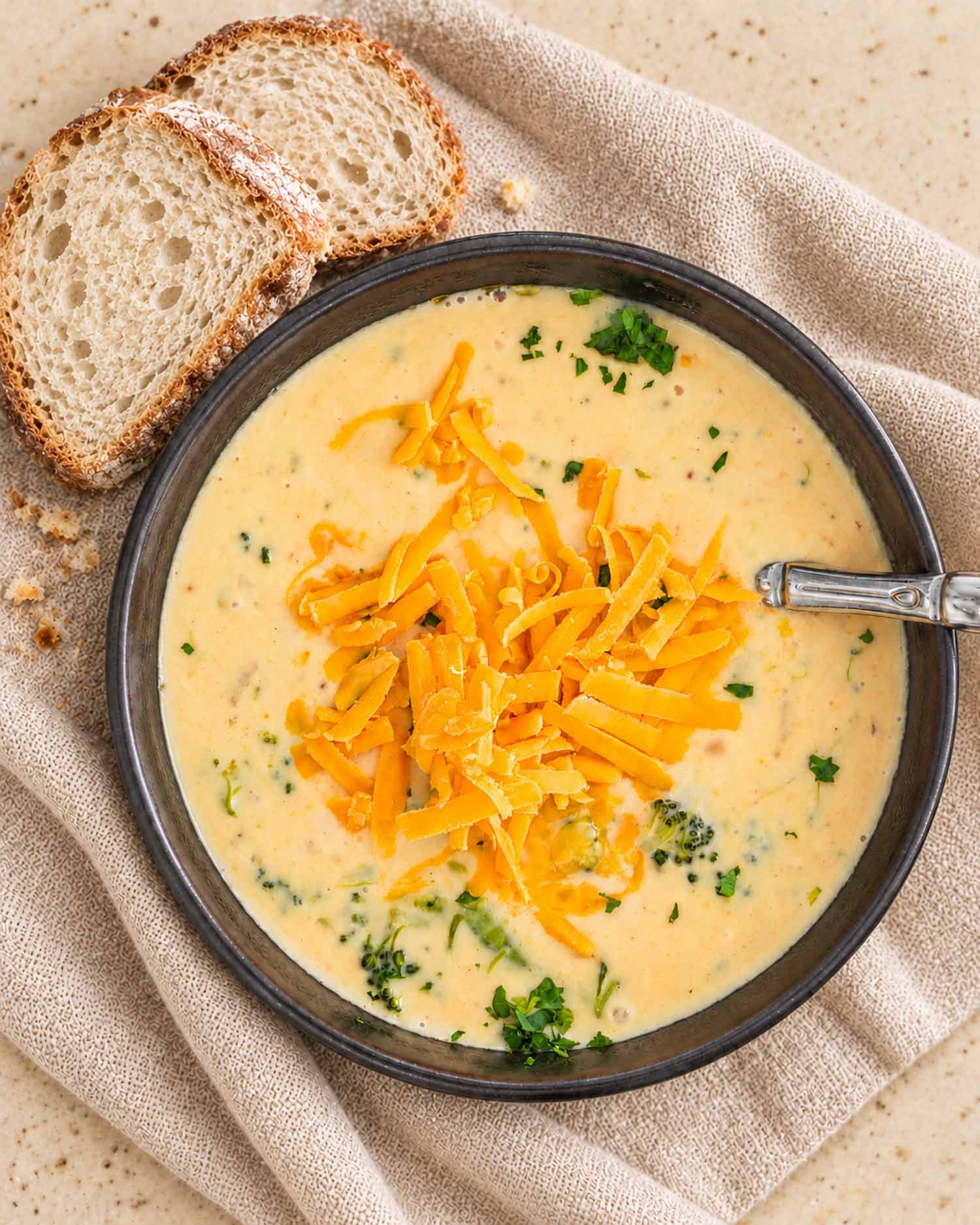 Overhead view of broccoli cheddar soup topped with shredded cheese and parsley, served beside crusty bread on beige linen.