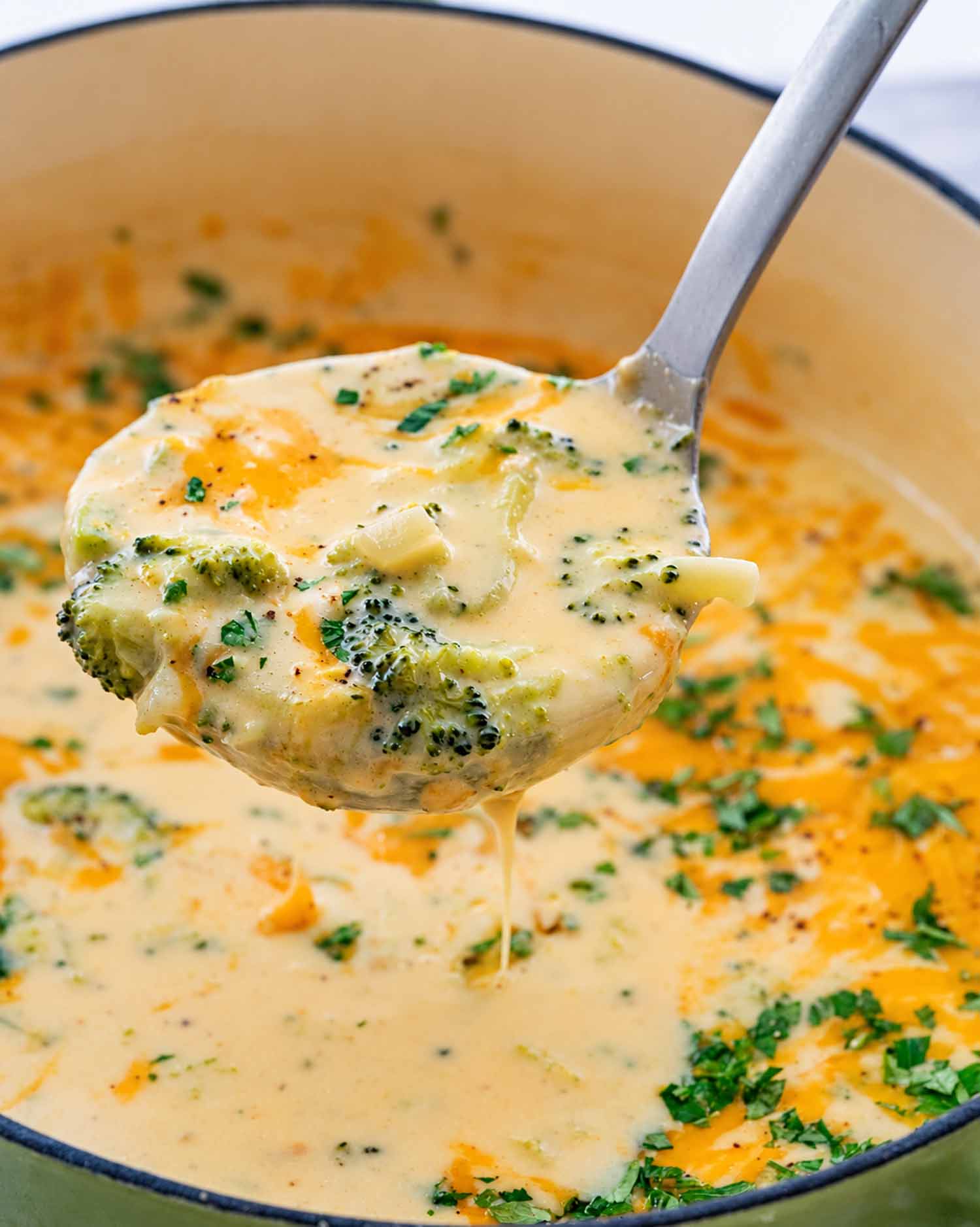 A metal ladle holds thick broccoli cheddar soup above the pot, with broccoli florets and a thin drip beneath.