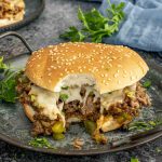 Philly Cheesesteak Sloppy Joe with a bite taken out, showing the cheesy, saucy beef filling with peppers and onions on a soft sesame bun.
