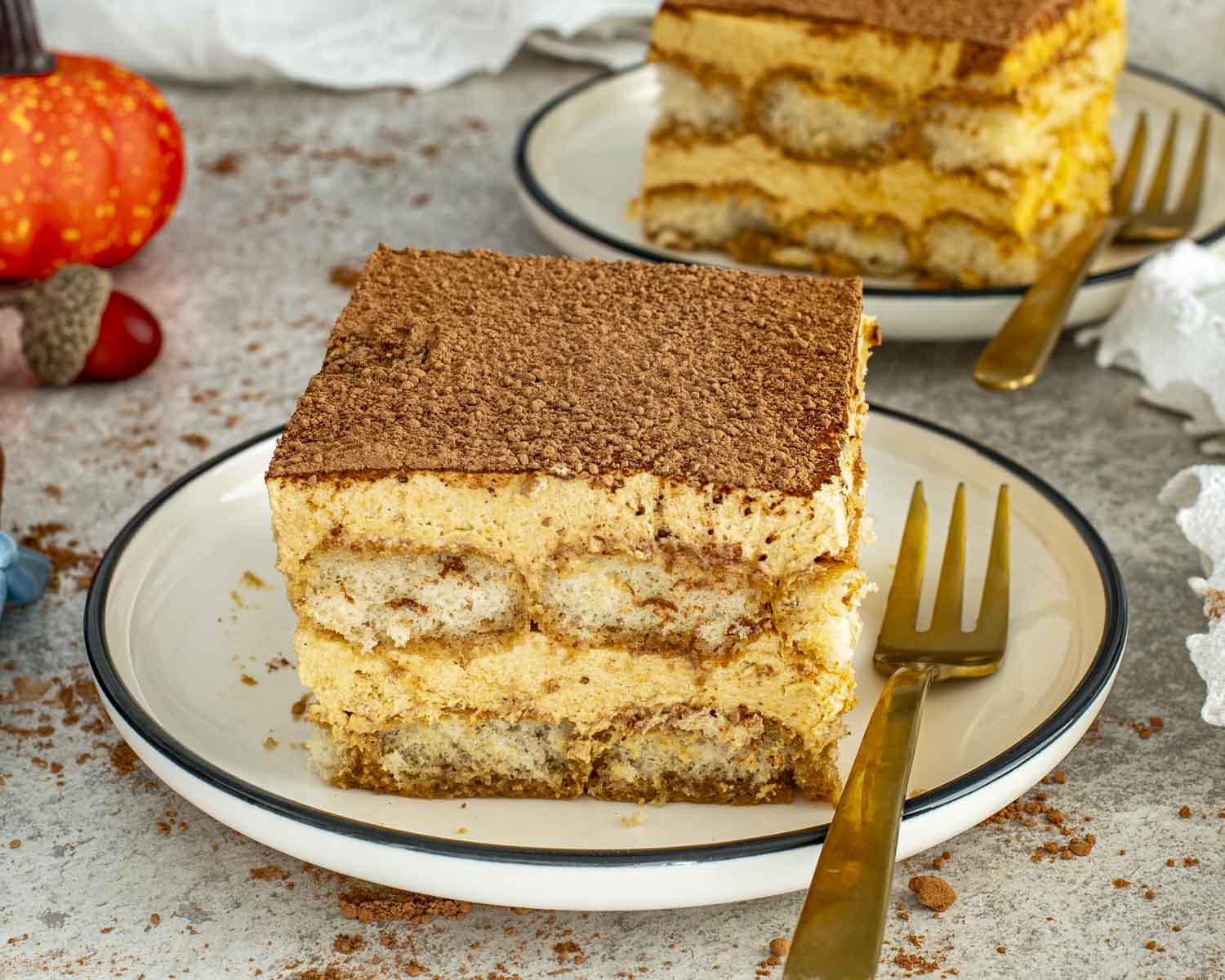 Two slices of pumpkin tiramisu on white plates, layers of ladyfingers and pumpkin cream topped with cocoa powder.