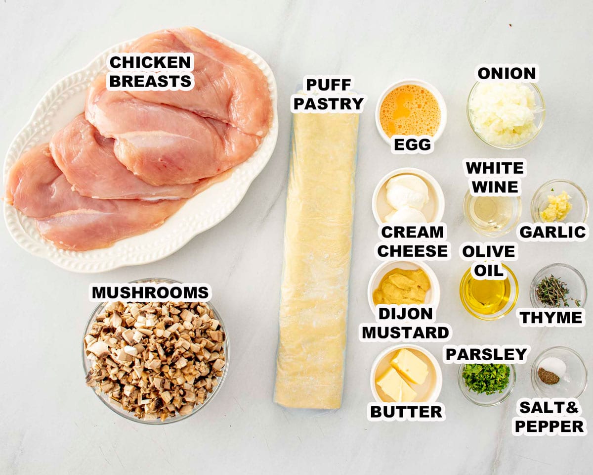 Overhead view of Chicken Wellington ingredients including chicken breasts, puff pastry, mushrooms, Dijon mustard, herbs, butter, and seasonings.