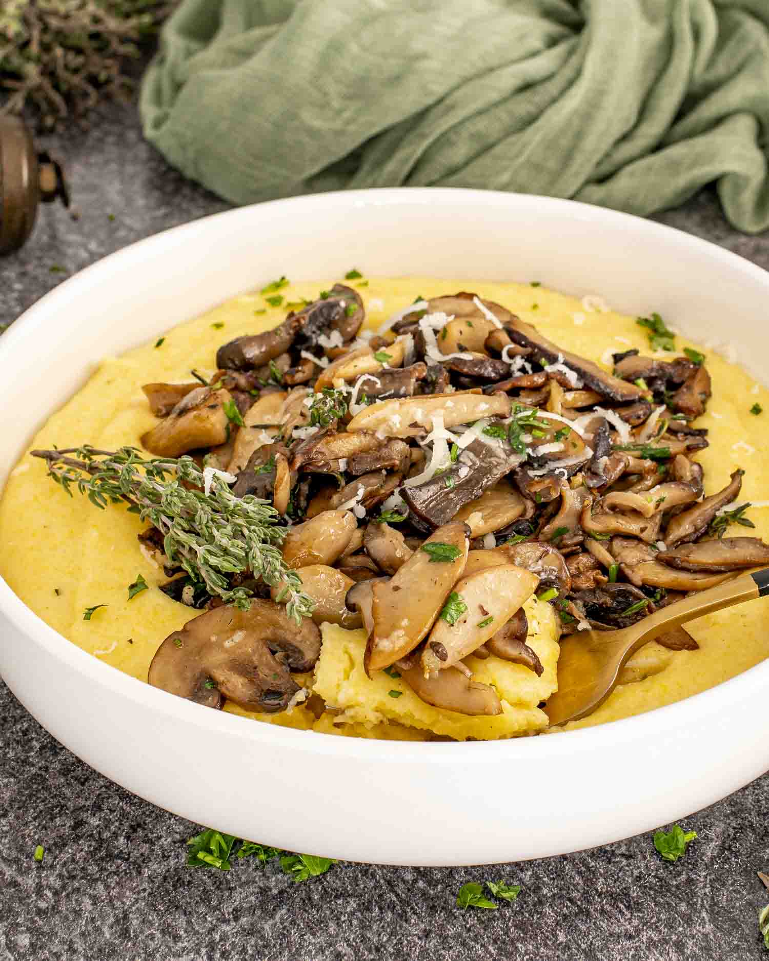 Creamy yellow polenta in a white bowl topped with sautéed mushrooms, thyme sprigs, and freshly grated Parmesan cheese.
