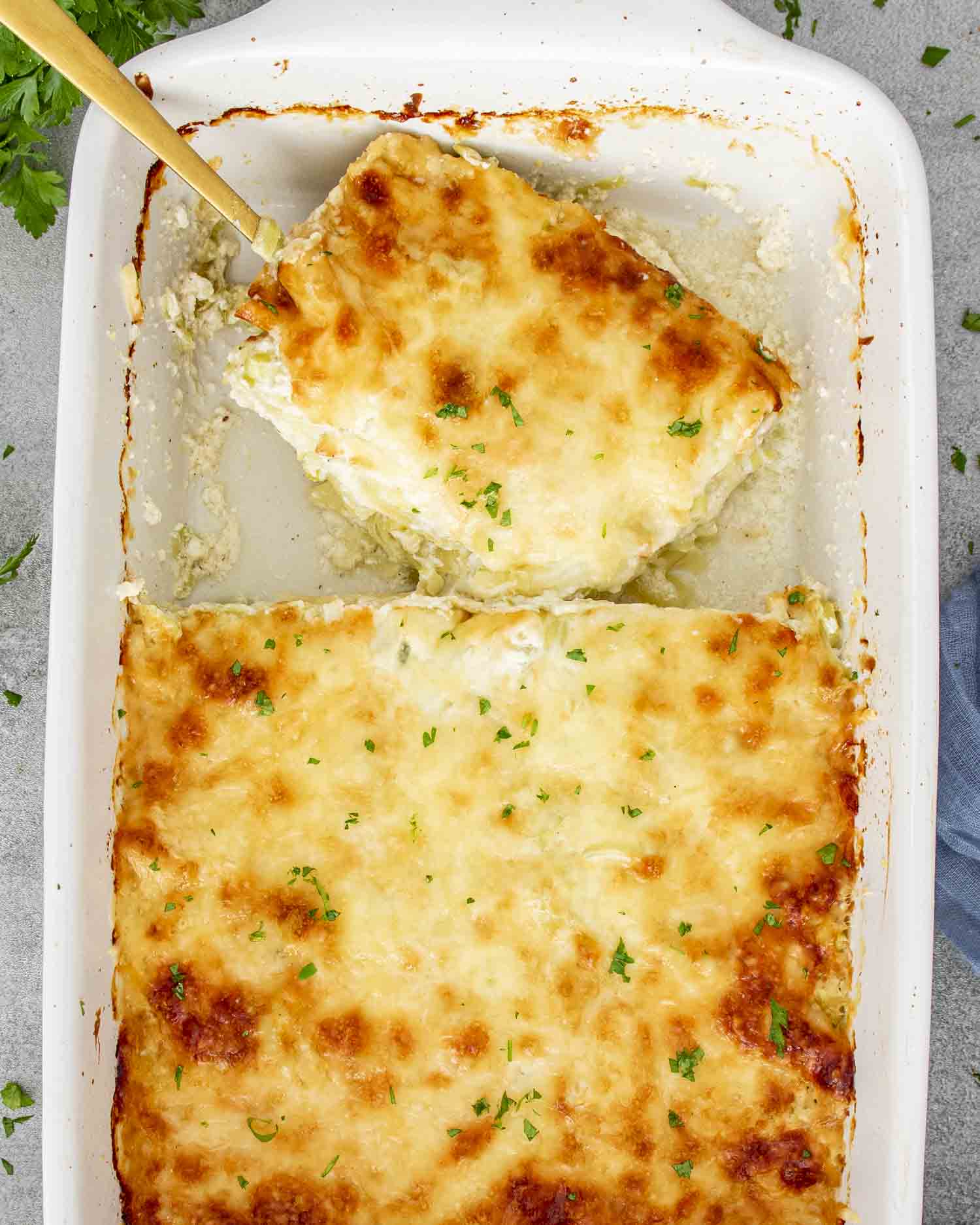 Gruyère and leek scalloped potatoes in white baking dish, golden cheese crust, slice being served to reveal creamy layers.