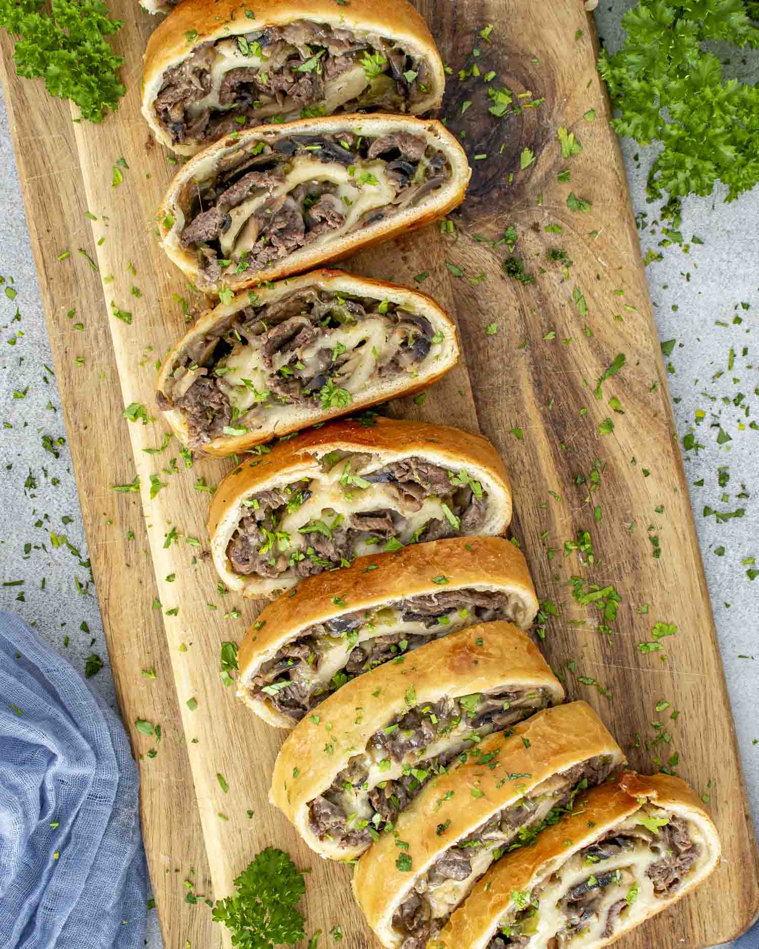 Top down view of sliced Philly cheesesteak stromboli arranged on a wooden board with parsley scattered around.