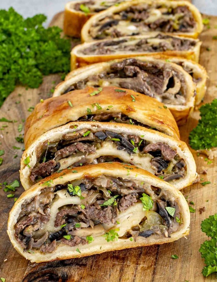 Warm Philly cheesesteak stromboli slices on a wooden board filled with ribeye, onions, peppers, mushrooms, and melted cheese.