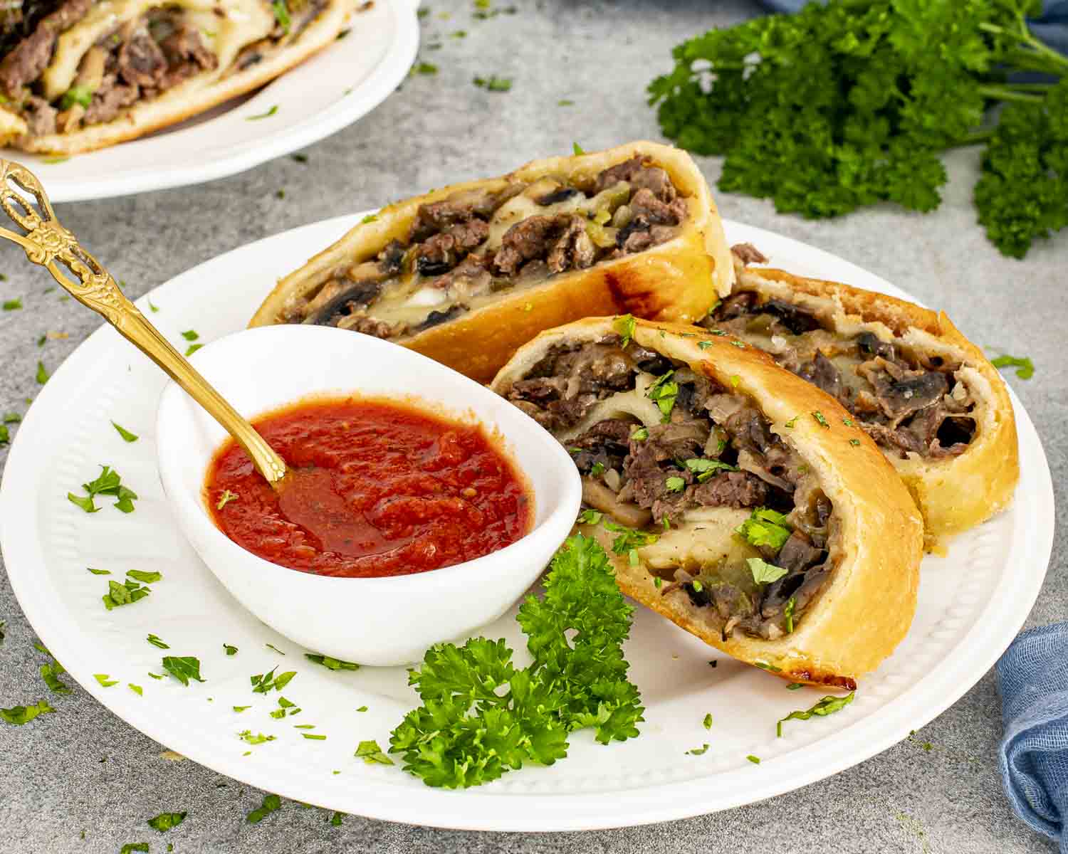 Three slices of Philly cheesesteak stromboli on a white plate with marinara dipping sauce and fresh chopped parsley.
