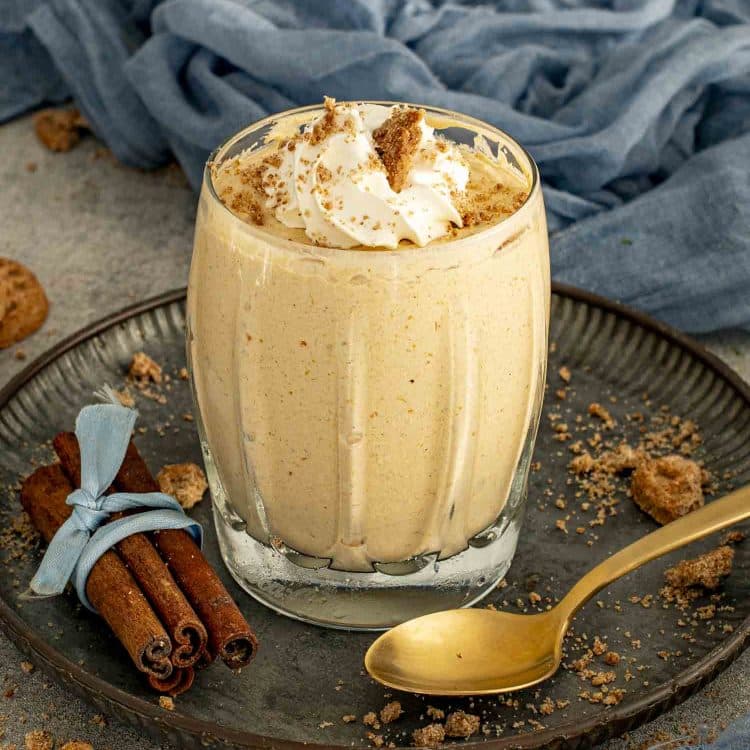Pumpkin mousse in a glass topped with whipped cream and cookie crumbs served on a plate with cinnamon sticks beside it.