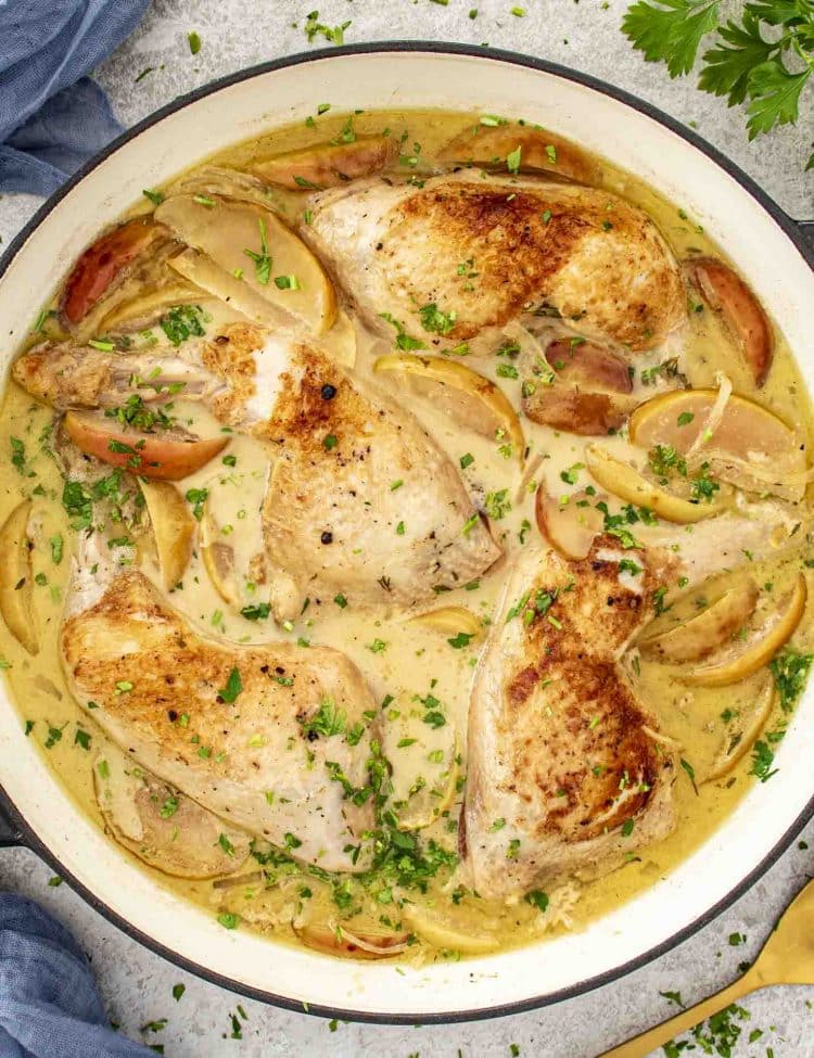Overhead shot of Chicken Normandy simmering in creamy apple cider sauce with apples, onions, thyme, and golden seared chicken legs.