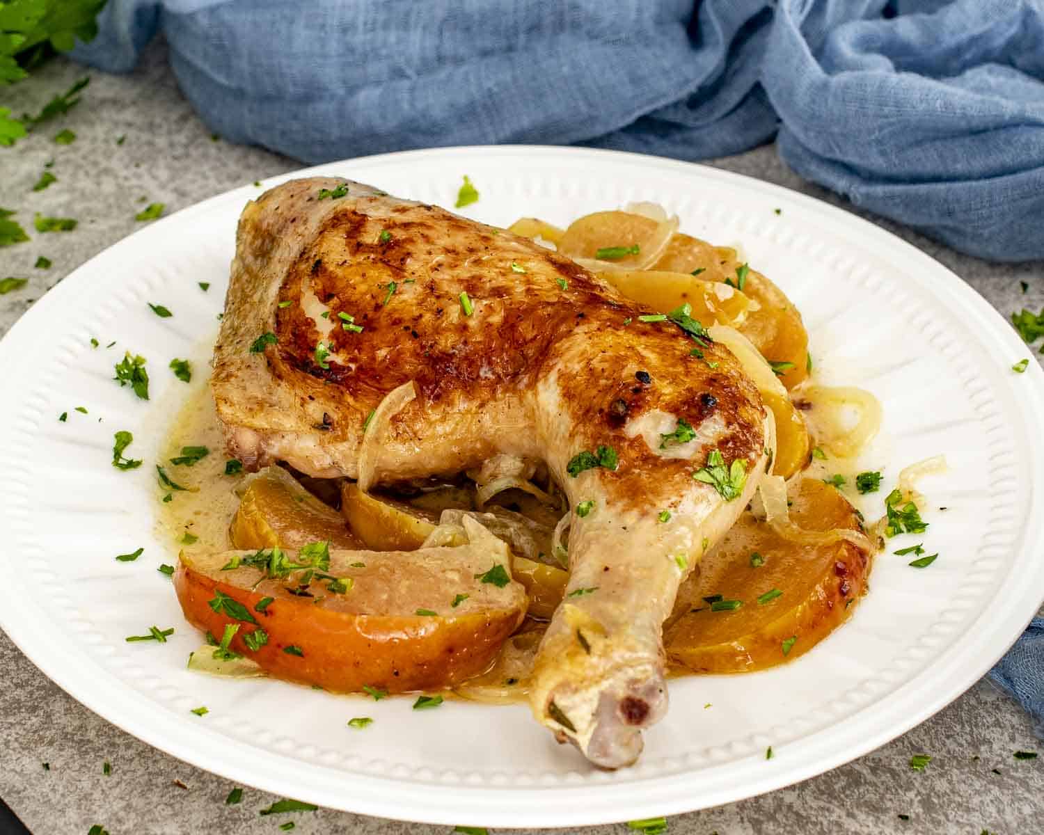Single chicken leg served with braised apples, onions, and creamy Normandy sauce, garnished with fresh parsley on a white plate.