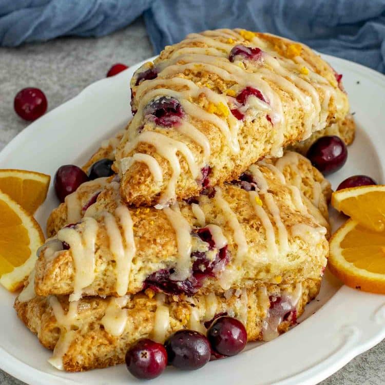 A plate stacked with freshly baked cranberry orange scones topped with sweet orange glaze and garnished with sliced oranges and cranberries.
