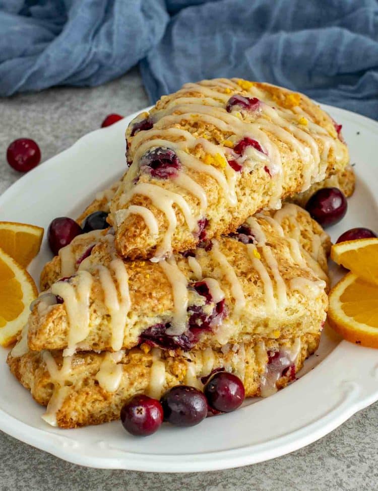 A plate stacked with freshly baked cranberry orange scones topped with sweet orange glaze and garnished with sliced oranges and cranberries.