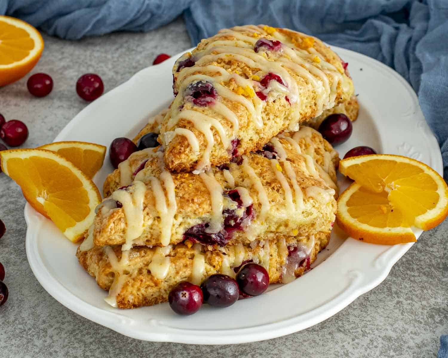 Stacked cranberry orange scones on a serving platter with fresh orange wedges and cranberries adding bright color and texture.