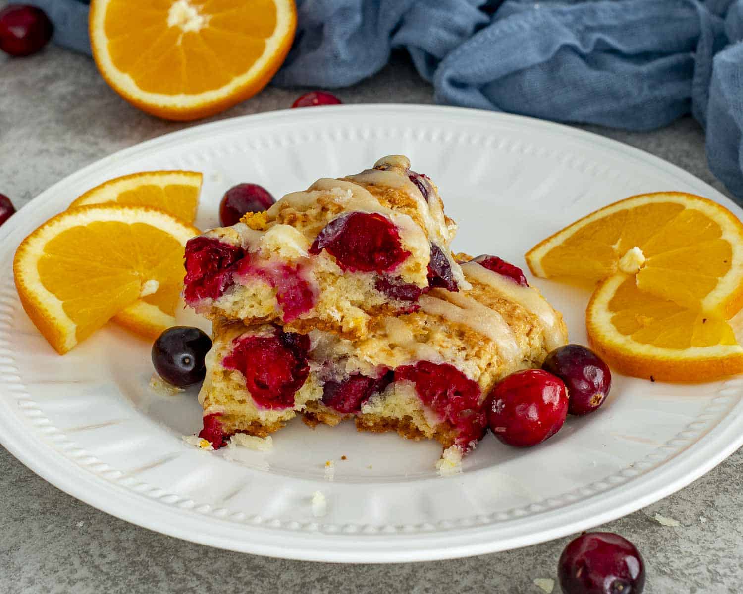 Close up of cranberry orange scones on a white plate, highlighting juicy cranberries, tender crumb, and glossy orange glaze drizzle.