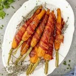 Large platter of maple bacon roasted carrots with fresh thyme, highlighting the golden caramelized glaze and tightly wrapped crispy bacon.