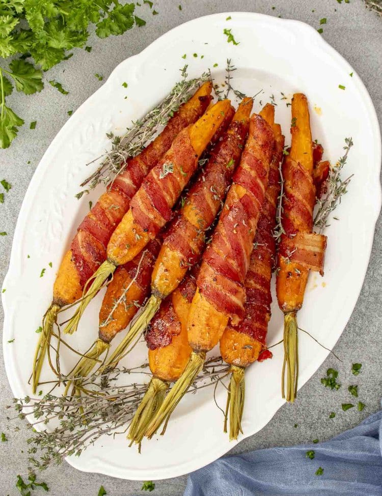 Large platter of maple bacon roasted carrots with fresh thyme, highlighting the golden caramelized glaze and tightly wrapped crispy bacon.