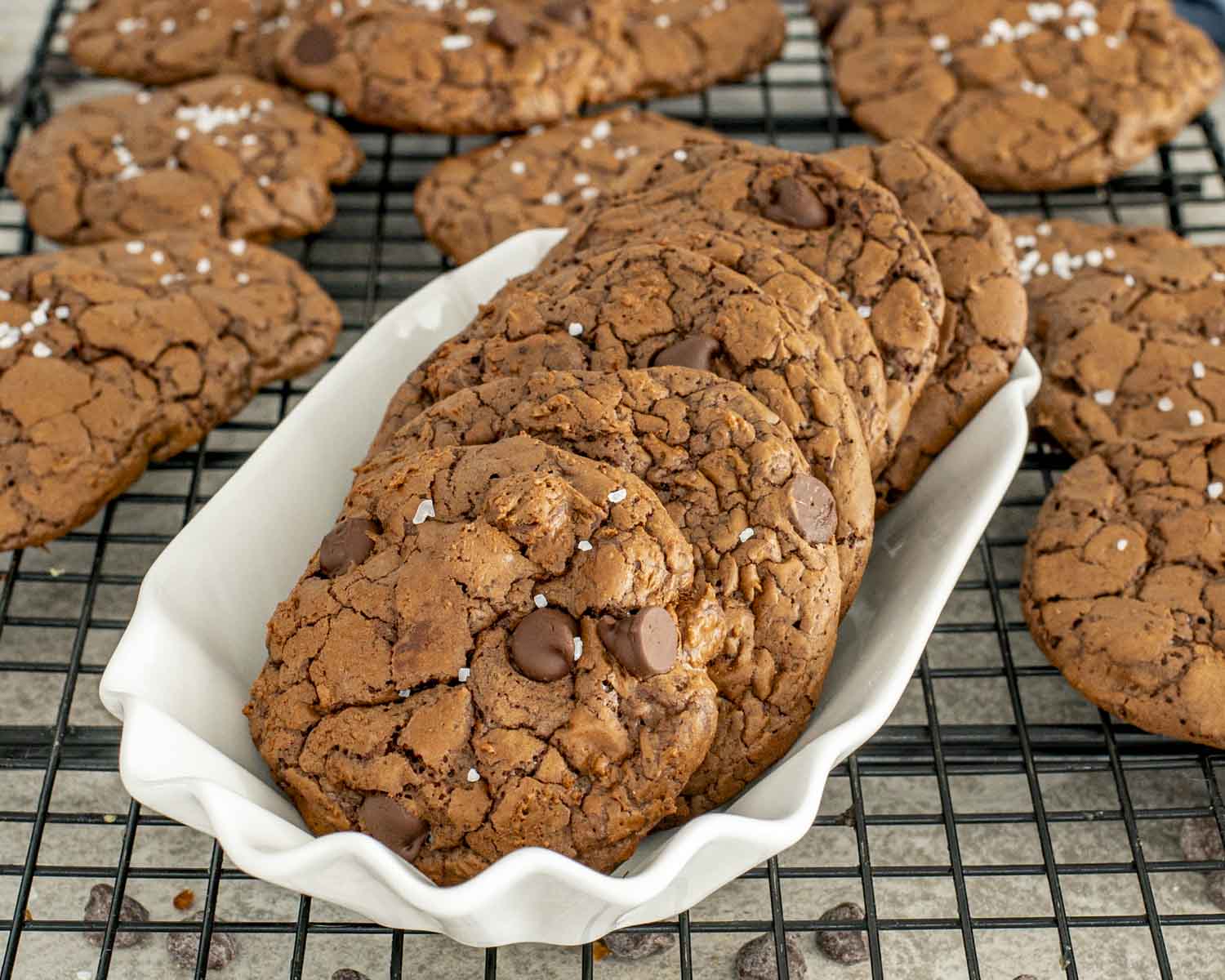 Mocha fudge cookies stacked in a white dish on a cooling rack, featuring crackled tops, chocolate chips, and flaky salt.
