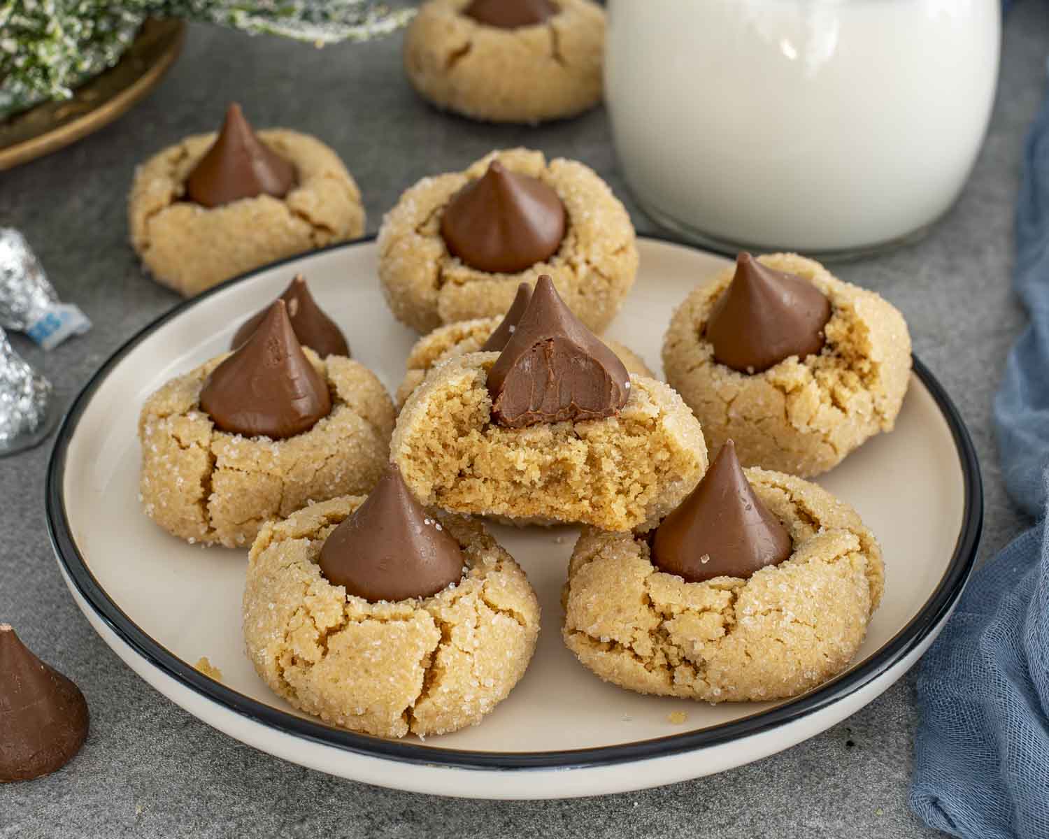 Freshly baked peanut butter blossom cookies arranged on a plate, each topped with a chocolate kiss, served beside a glass of milk.