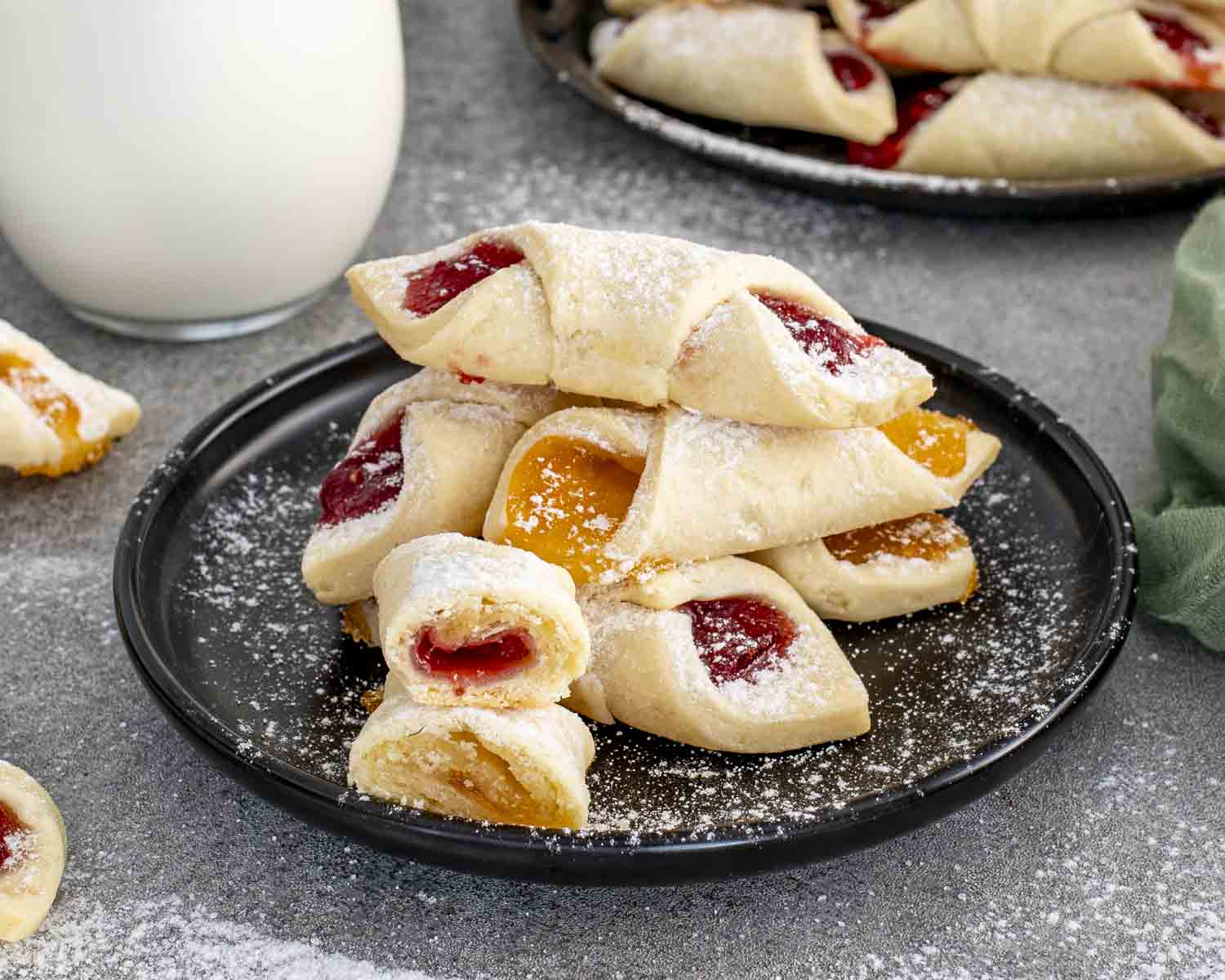 Stack of homemade Polish Kolaczki filled with apricot and raspberry preserves, dusted with powdered sugar on a dark plate beside milk.