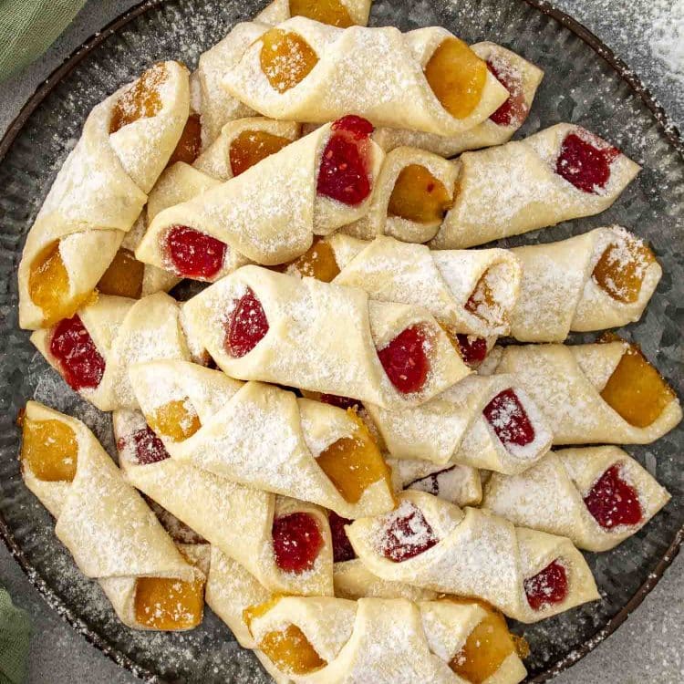 Close-up plate of soft Polish Kolaczki cookies showing apricot and raspberry fillings, lightly powdered with sugar for a festive finish.