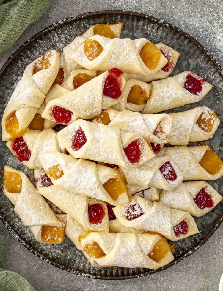 Close-up plate of soft Polish Kolaczki cookies showing apricot and raspberry fillings, lightly powdered with sugar for a festive finish.