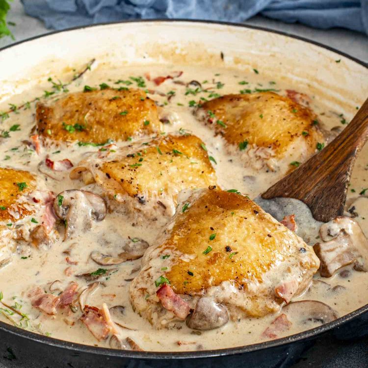 Coq au Riesling simmering in a skillet with golden chicken thighs, mushrooms, bacon, thyme, and creamy white wine sauce.