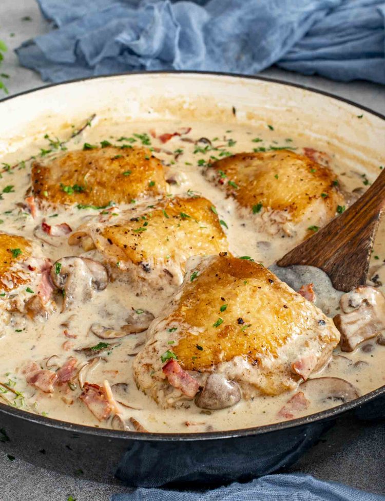 Coq au Riesling simmering in a skillet with golden chicken thighs, mushrooms, bacon, thyme, and creamy white wine sauce.