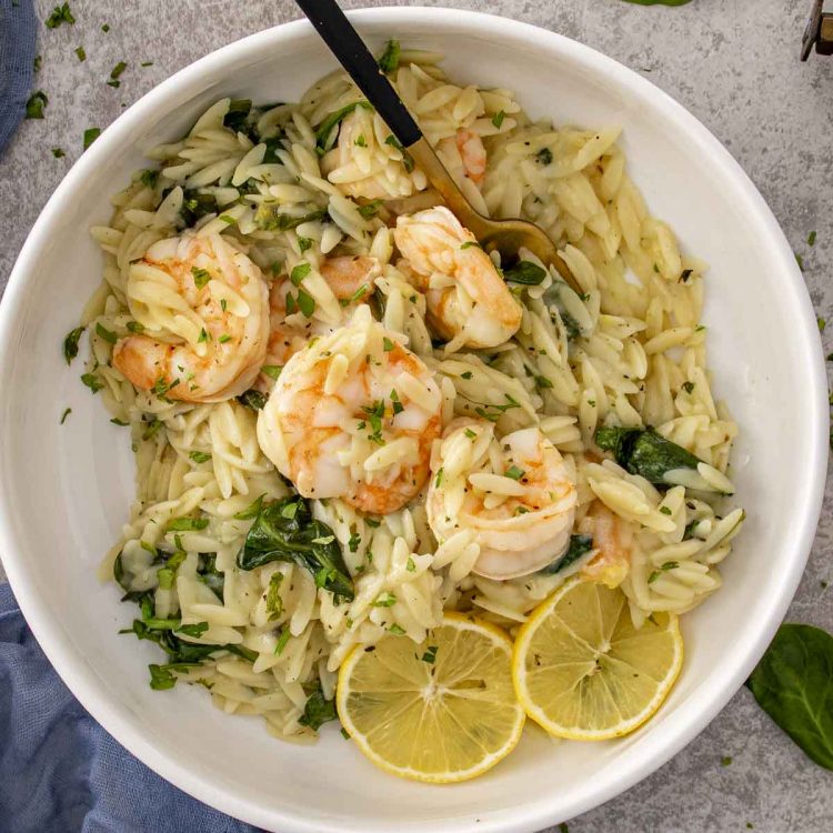 Overhead view of lemon garlic shrimp orzo topped with juicy shrimp, fresh herbs, and lemon slices in a white bowl.