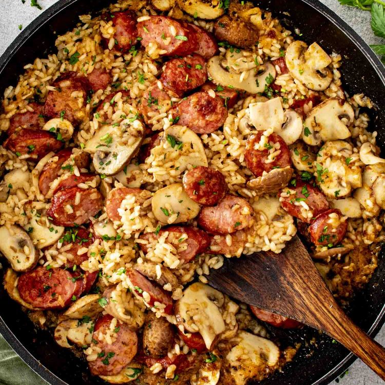 Overhead view of skillet sausage and mushroom rice cooking in a pan with smoked sausage, mushrooms, rice, and fresh parsley.