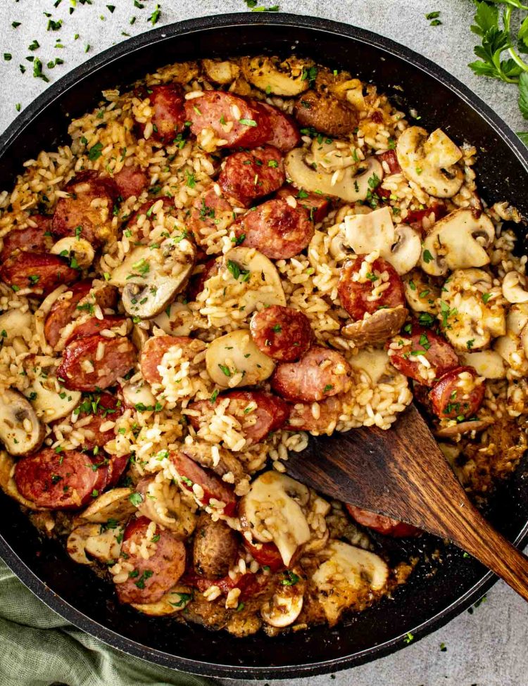 Overhead view of skillet sausage and mushroom rice cooking in a pan with smoked sausage, mushrooms, rice, and fresh parsley.