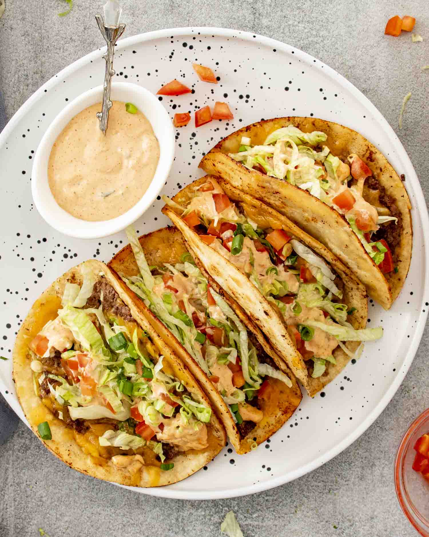 Smashed burger tacos topped with lettuce, tomatoes, green onions, and creamy Big Mac style sauce on plate.