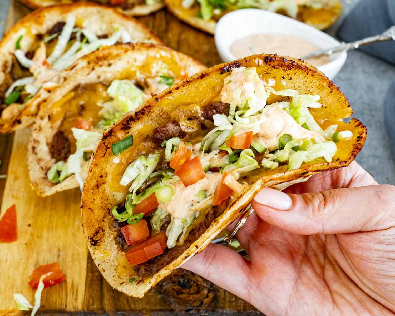 Hand holding crispy smashed burger taco showing juicy beef, melted cheese, lettuce, tomatoes, and creamy burger sauce.