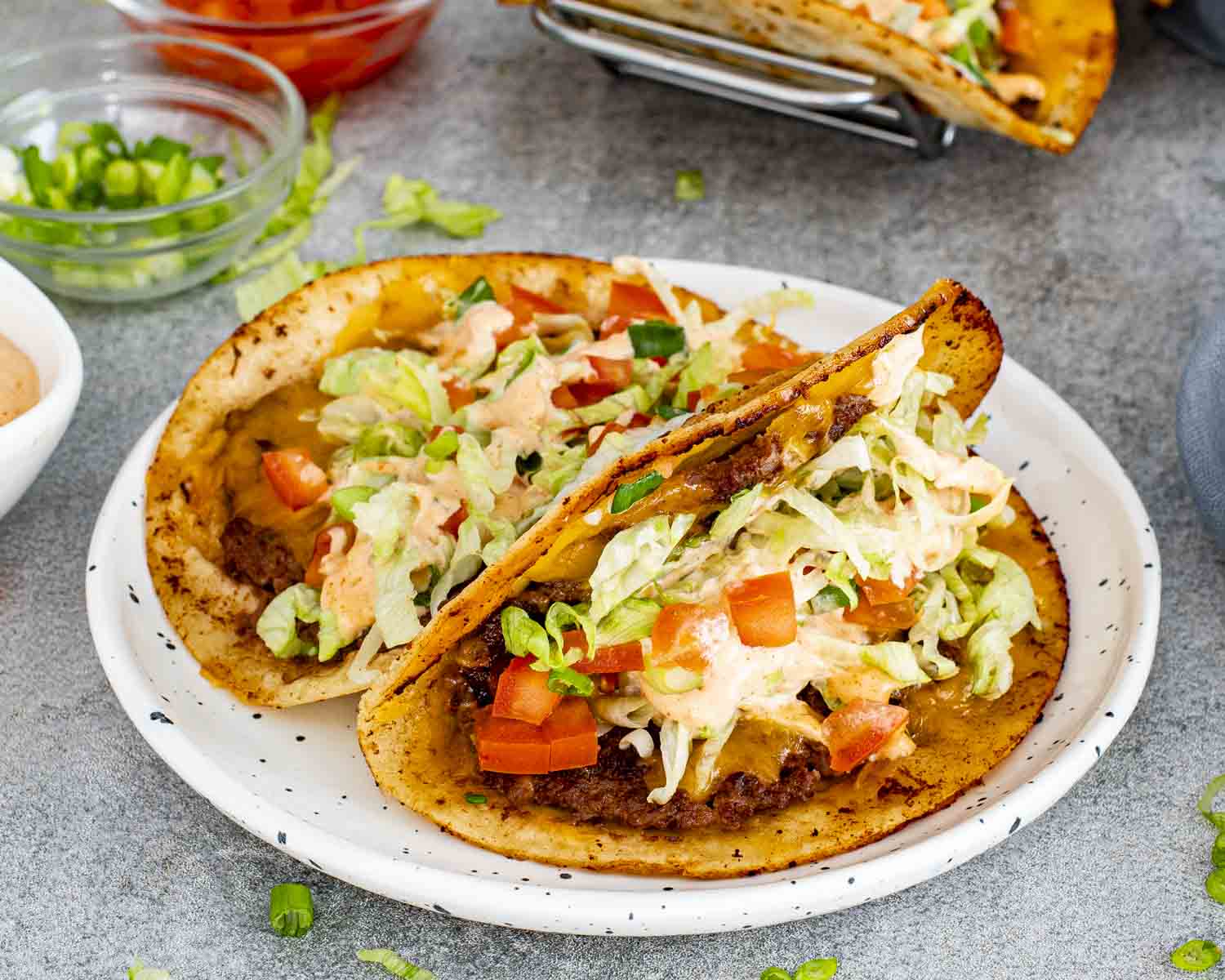 Golden smashed burger tacos served on plate, filled with seasoned beef, cheddar cheese, lettuce, tomatoes, and sauce.