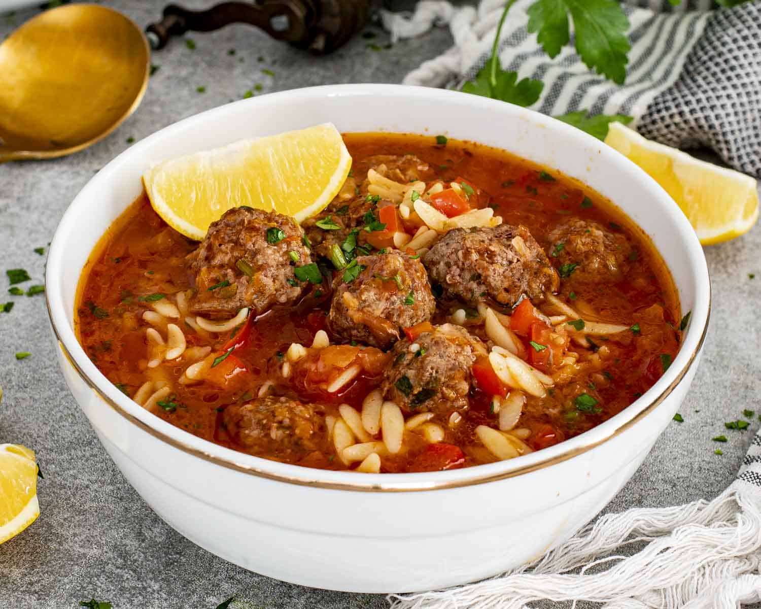 Turkish meatball soup served in white bowl with orzo, beef meatballs, tomato broth, parsley, and lemon on side.