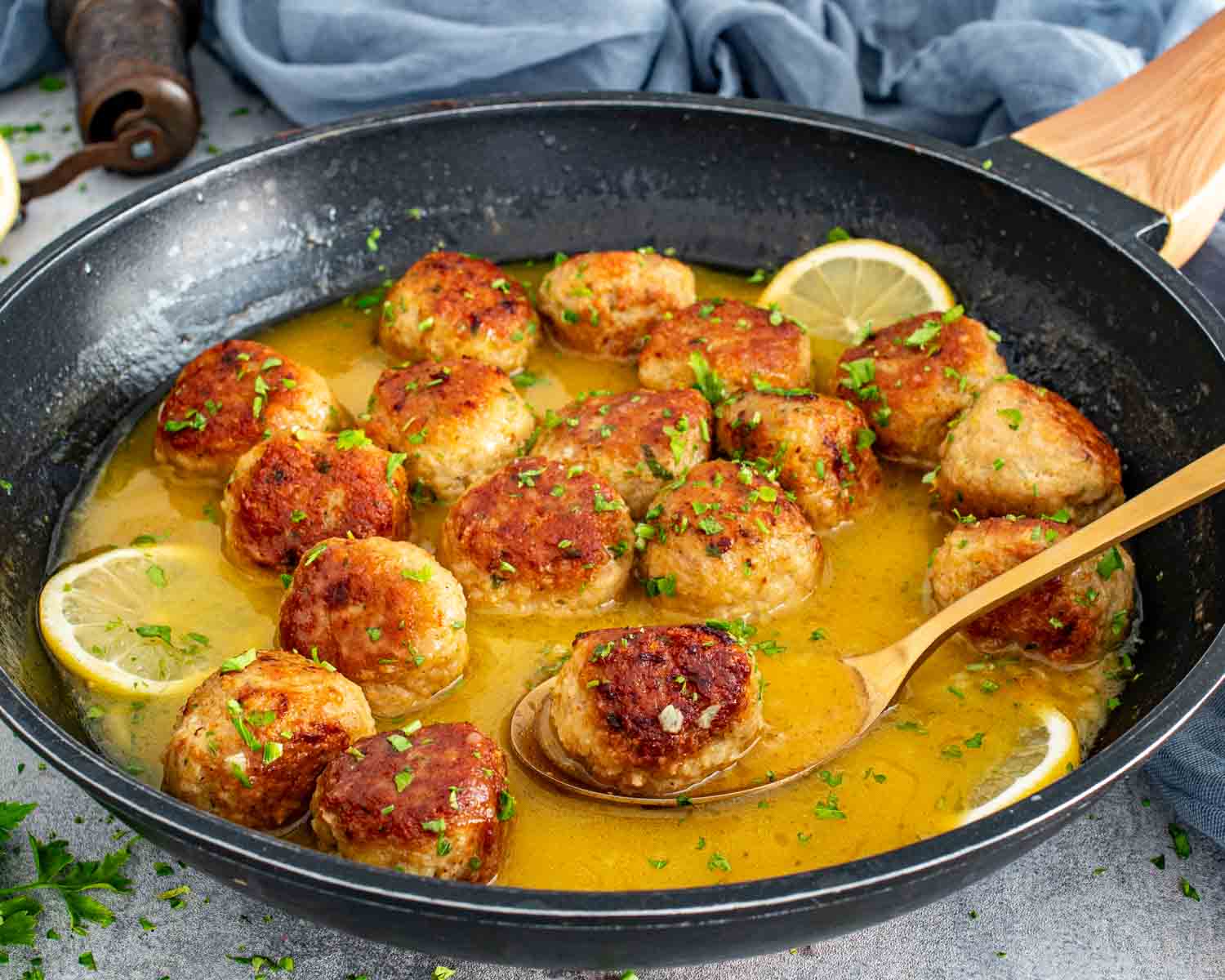 Chicken Meatballs in Lemon Butter Sauce cooking in a skillet with a spoon lifting a browned meatball.