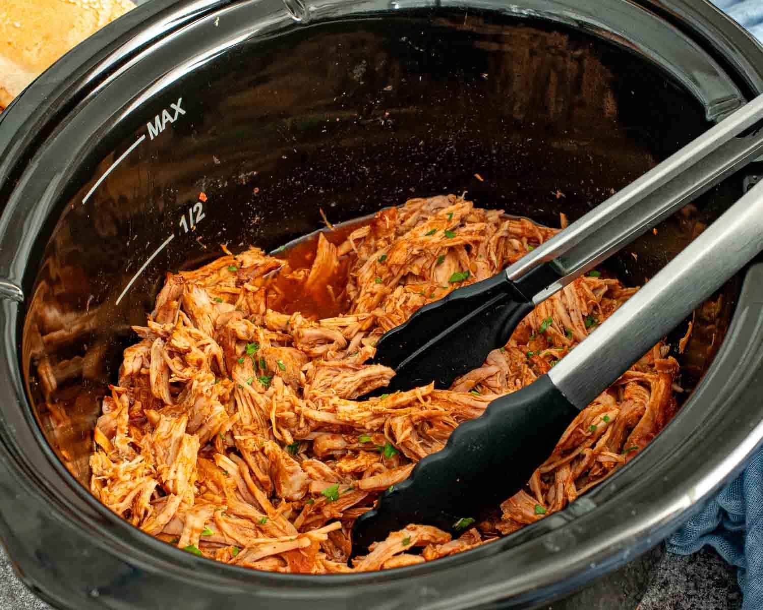 Slow cooker filled with shredded BBQ pulled pork, mixed and ready for serving using kitchen tongs.