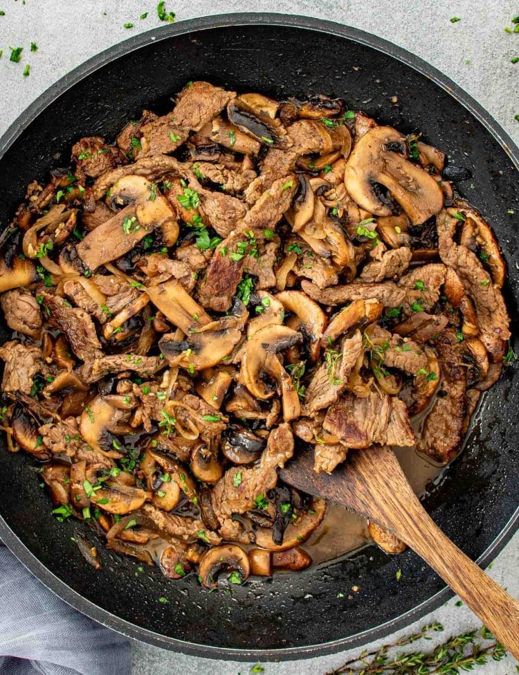 Steak and mushrooms cooking together in a large skillet with butter, onions, thyme, and savory sauce.