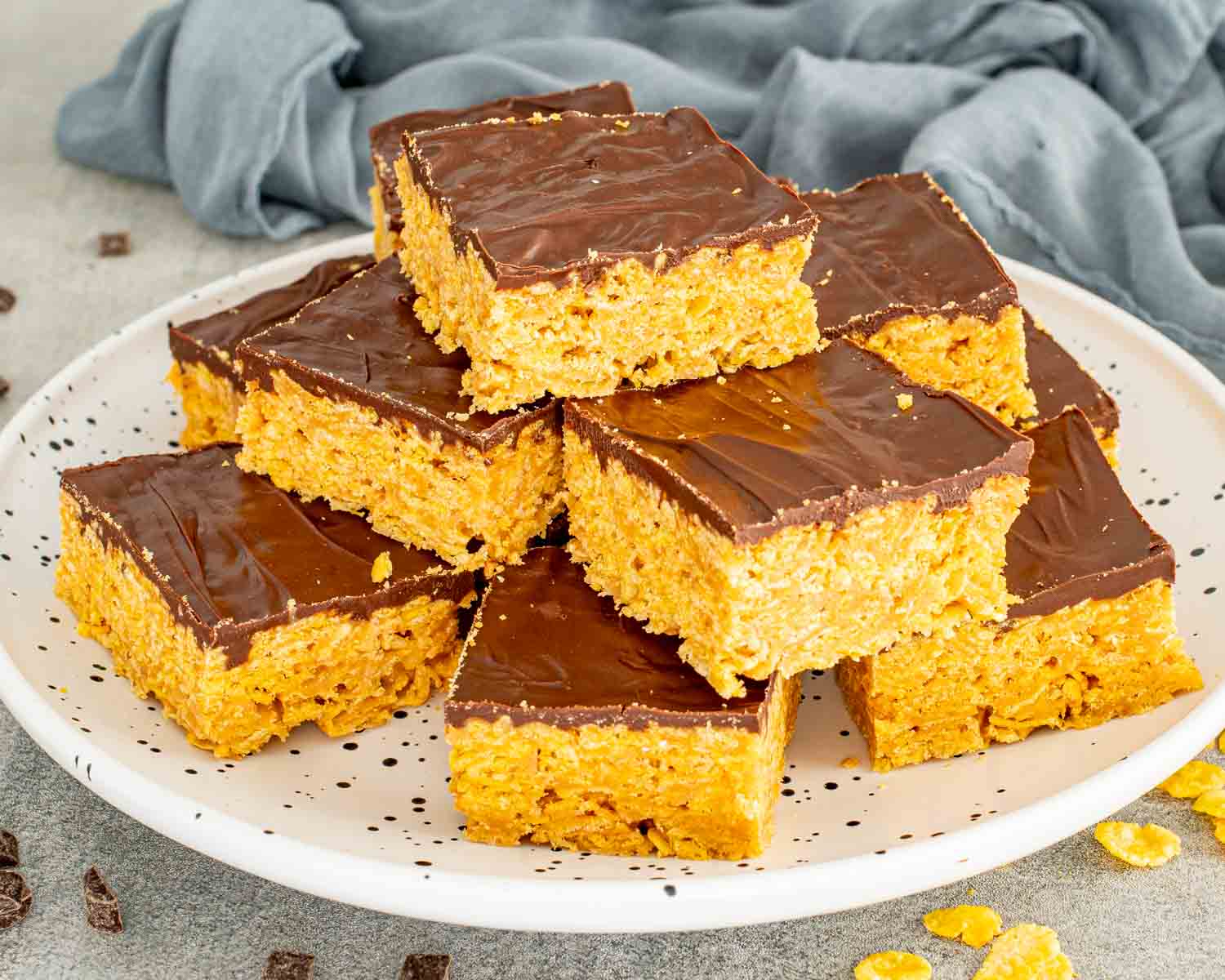No bake Butterfinger bars stacked on a white plate with crisp cornflake peanut butter base and chocolate topping.