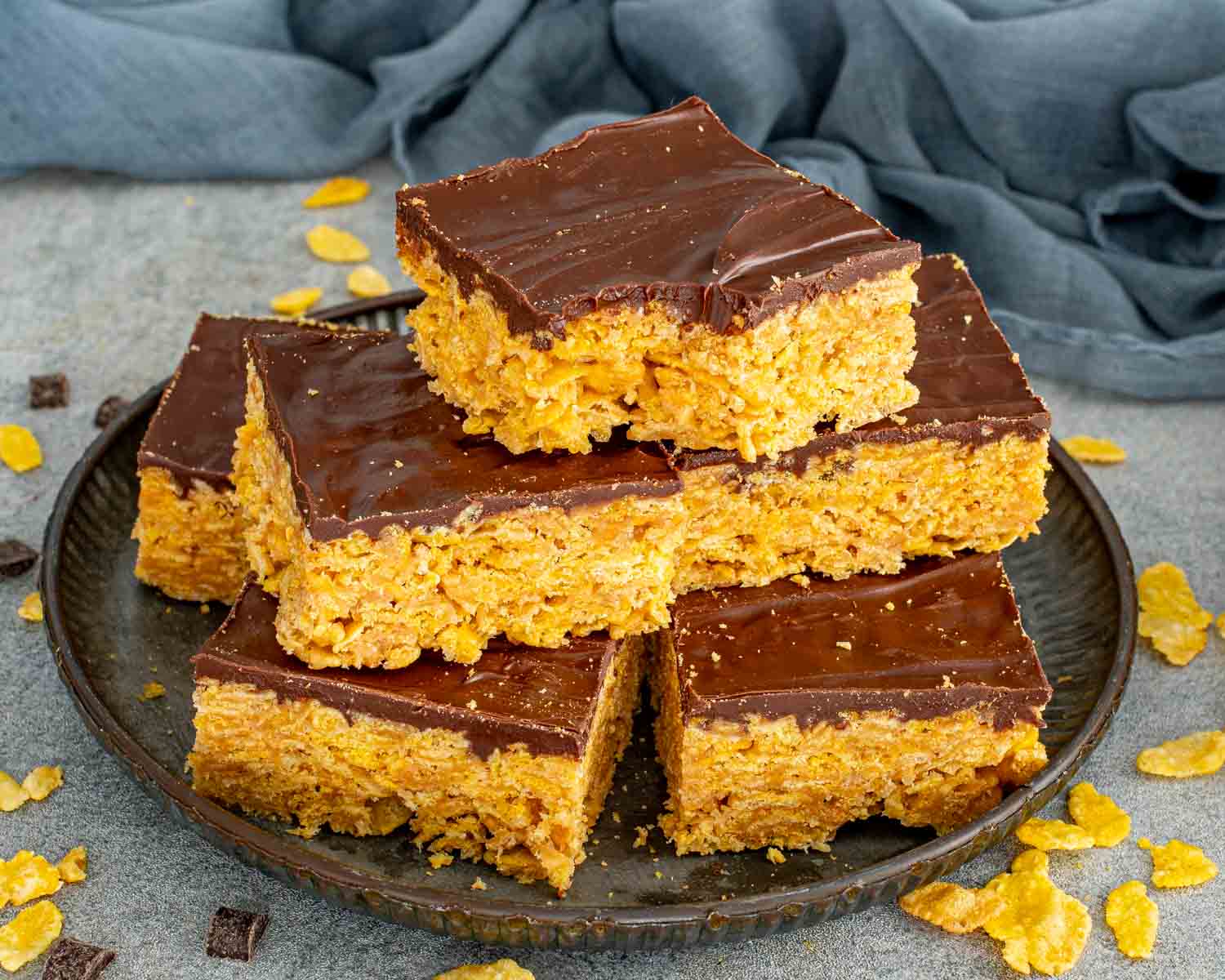 Homemade Butterfinger bars stacked on a dark plate with crunchy cornflake peanut butter base and smooth chocolate layer.