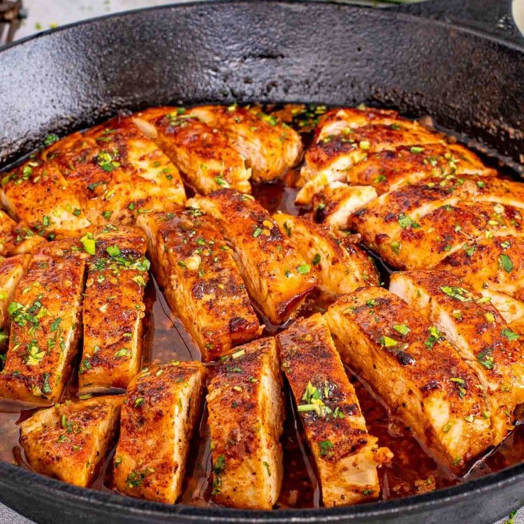 Close-up of juicy chicken breasts simmering in a cast iron skillet with glossy garlic butter sauce and fresh parsley.