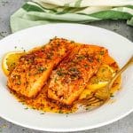 Two salmon fillets served with lemon slices and spooned cowboy butter sauce on white dinner plate.